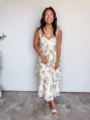 A full-length photograph of a smiling woman with dark wavy hair looking upward against a textured white wall. She is wearing a sleeveless tiered midi dress made from a 75% polyester and 25% nylon blend, featuring a muted taupe and beige floral print on a cream base, a fitted bustier bodice with a sweetheart neckline, ruffled shoulder straps, and a multi-tiered flared midi skirt, paired with cream open-toe slide heels and styled with a gold pendant necklace and gold beaded bracelets.