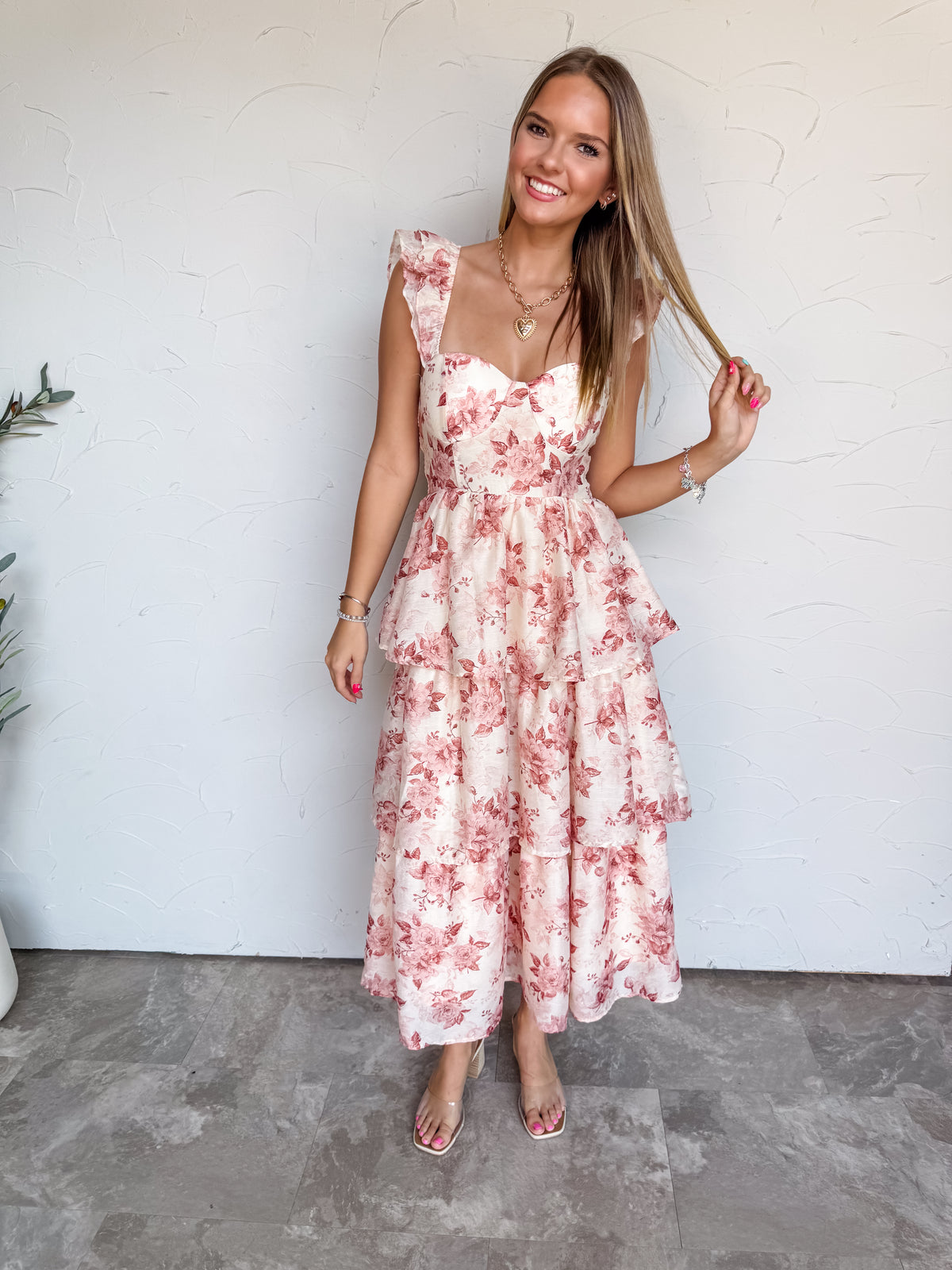 A full-length photograph of a smiling young woman with long, straight blonde-highlighted hair touching a strand of her hair against a textured white wall. She is wearing a sleeveless tiered midi dress made from a 75% polyester and 25% nylon blend, featuring a muted blush pink and mauve floral print on a cream base, a fitted bustier bodice with a sweetheart neckline, ruffled shoulder straps, and a multi-tiered flared midi skirt, paired with clear strap heels and styled with a gold heart pendant necklace and 