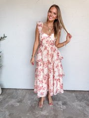 A full-length photograph of a smiling young woman with long, straight blonde-highlighted hair touching a strand of her hair against a textured white wall. She is wearing a sleeveless tiered midi dress made from a 75% polyester and 25% nylon blend, featuring a muted blush pink and mauve floral print on a cream base, a fitted bustier bodice with a sweetheart neckline, ruffled shoulder straps, and a multi-tiered flared midi skirt, paired with clear strap heels and styled with a gold heart pendant necklace and 