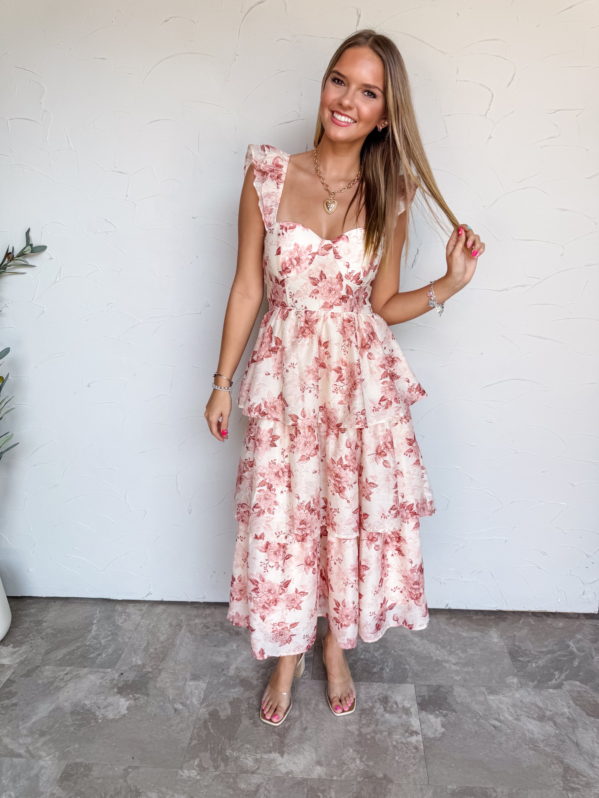 A full-length photograph of a smiling young woman with long, straight blonde-highlighted hair touching a strand of her hair against a textured white wall. She is wearing a sleeveless tiered midi dress made from a 75% polyester and 25% nylon blend, featuring a muted blush pink and mauve floral print on a cream base, a fitted bustier bodice with a sweetheart neckline, ruffled shoulder straps, and a multi-tiered flared midi skirt, paired with clear strap heels and styled with a gold heart pendant necklace and 