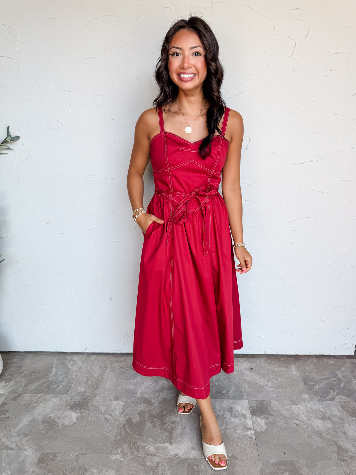 A full-length photograph of a smiling woman with dark wavy hair standing with one hand in her pocket against a textured white wall. She is wearing a sleeveless deep red midi dress made from a 65% cotton, 32% nylon, and 3% spandex blend, featuring wide shoulder straps, white contrast topstitching along the fitted bustier bodice and skirt seams, a matching self-tie belt at the waist, side pockets, and a flared midi skirt, paired with cream open-toe slide heels and styled with a gold coin pendant necklace and 