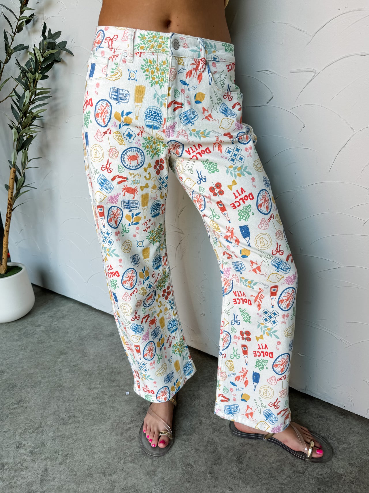 A close-up photograph focusing from the waist down of a woman wearing straight-leg white denim jeans featuring a colorful allover European vacation-themed print made from a 91% cotton, 6% polyester, and 3% spandex blend fabric against a textured white wall. The pants showcase whimsical illustrations of seafood, wine, and lemons, styled with flat clear strap sandals.