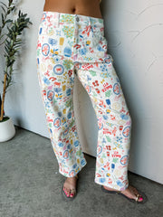 A close-up photograph focusing from the waist down of a woman wearing straight-leg white denim jeans featuring a colorful allover European vacation-themed print made from a 91% cotton, 6% polyester, and 3% spandex blend fabric against a textured white wall. The pants showcase whimsical illustrations of seafood, wine, and lemons, styled with flat clear strap sandals.