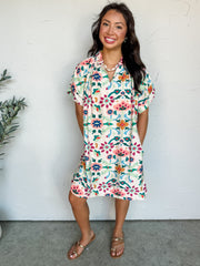 A full-length photograph of a smiling brunette woman posing with her hands in her pockets against a textured white wall. She is wearing a knee-length, short-sleeve shift dress made from 100% polyester fabric featuring a colorful pink, green, and blue abstract floral print on a cream background, styled with flat gold toe-loop sandals.