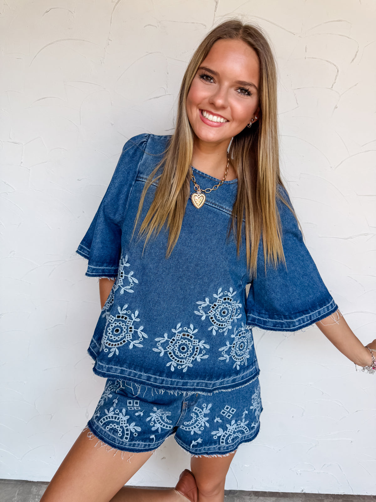 A full-length photograph of a smiling young woman with straight blonde-highlighted hair posing on one leg against a textured white wall. She is wearing a matching two-piece medium-wash denim set made from a 90% cotton and 10% polyester blend, featuring a short-sleeved denim top with light blue embroidered eyelet patterns and raw frayed flutter sleeves, paired with high-waisted denim shorts adorned with matching light blue eyelet embroidery and a frayed raw hem, styled with brown leather flats and a gold hea