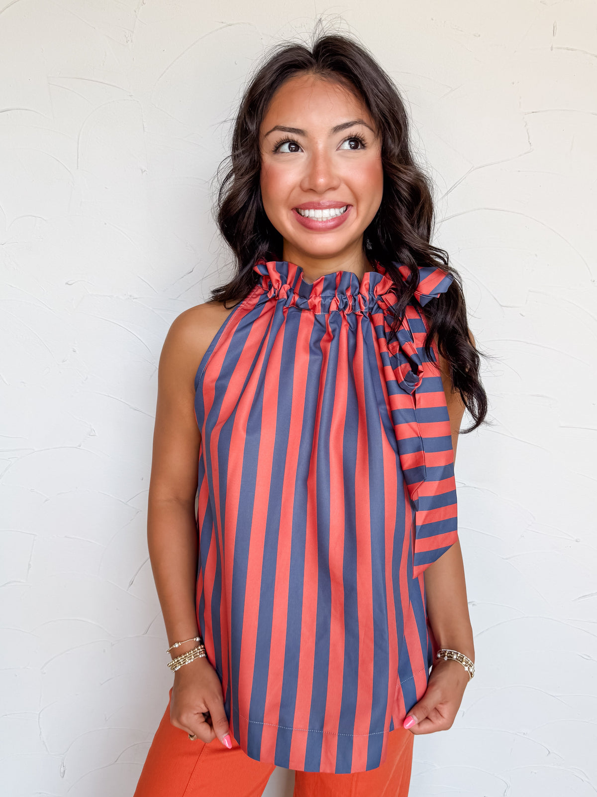 A waist-up photograph of a smiling woman with dark wavy hair looking upward and to the side against a textured white wall. She is wearing a sleeveless halter top made from 100% polyester, featuring a vertical stripe pattern in navy blue and burnt orange, a ruffled stand collar with a prominent side tie-bow accent, paired with burnt orange tailored pants and styled with gold beaded bracelets.