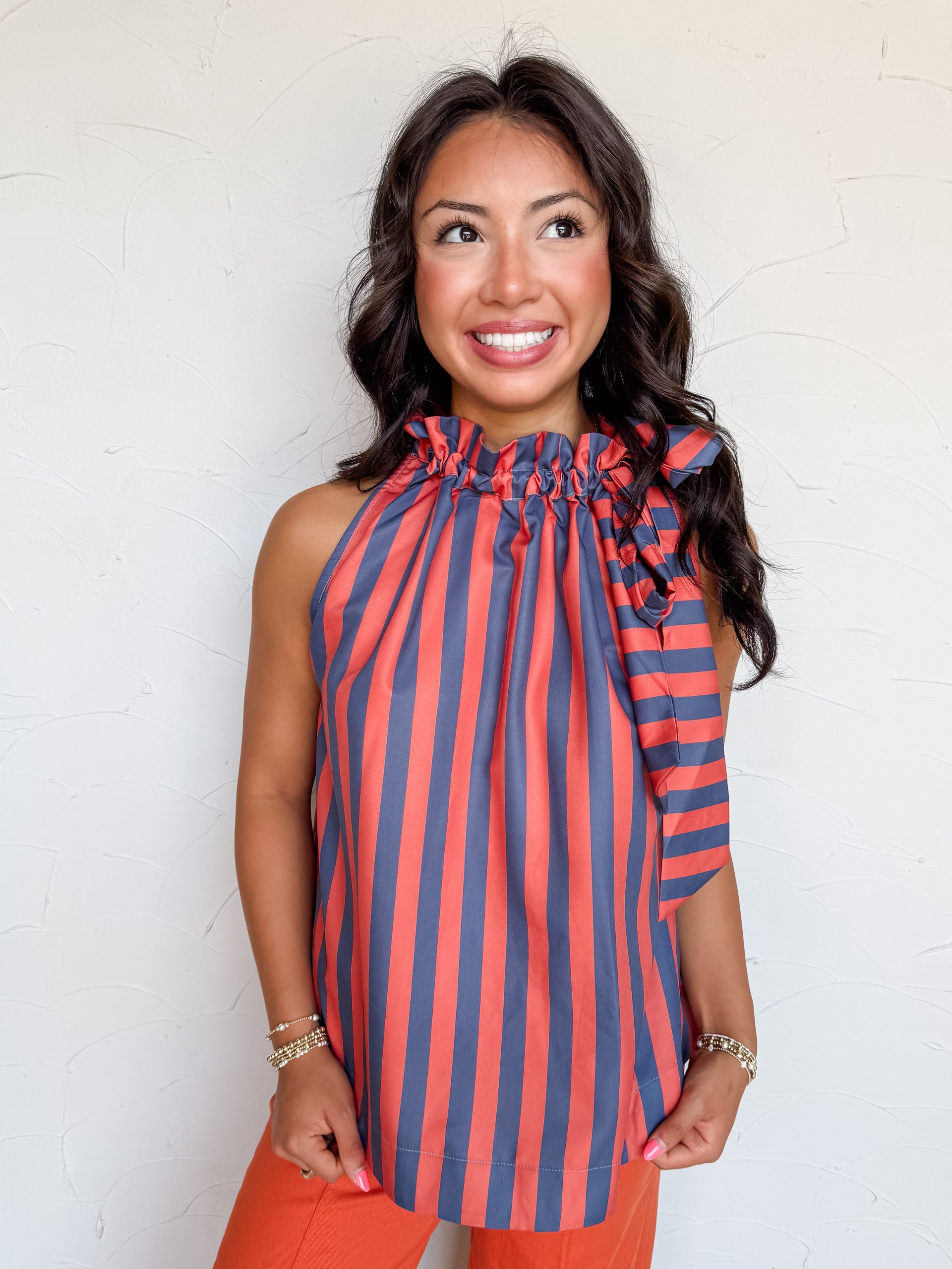 A waist-up photograph of a smiling woman with dark wavy hair looking upward and to the side against a textured white wall. She is wearing a sleeveless halter top made from 100% polyester, featuring a vertical stripe pattern in navy blue and burnt orange, a ruffled stand collar with a prominent side tie-bow accent, paired with burnt orange tailored pants and styled with gold beaded bracelets.