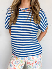 A close-up photograph from the chin down of a blonde woman posing with her hands behind her back against a textured white wall. She is wearing a relaxed short-sleeve top made from a textured 95% polyester and 5% spandex blend fabric featuring royal blue and white horizontal stripes, styled over white denim pants with colorful European illustrations.
