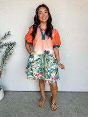 A full-length photograph of a smiling brunette woman posing with one hand in her pocket against a textured white wall. She is wearing a short puff-sleeve shift dress made from 100% cotton fabric featuring an orange-to-white ombre top and a detailed tropical coastal village print along the skirt, styled with contrast navy stitched trim on the V-neckline and sleeve cuffs, and flat gold toe-loop sandals.