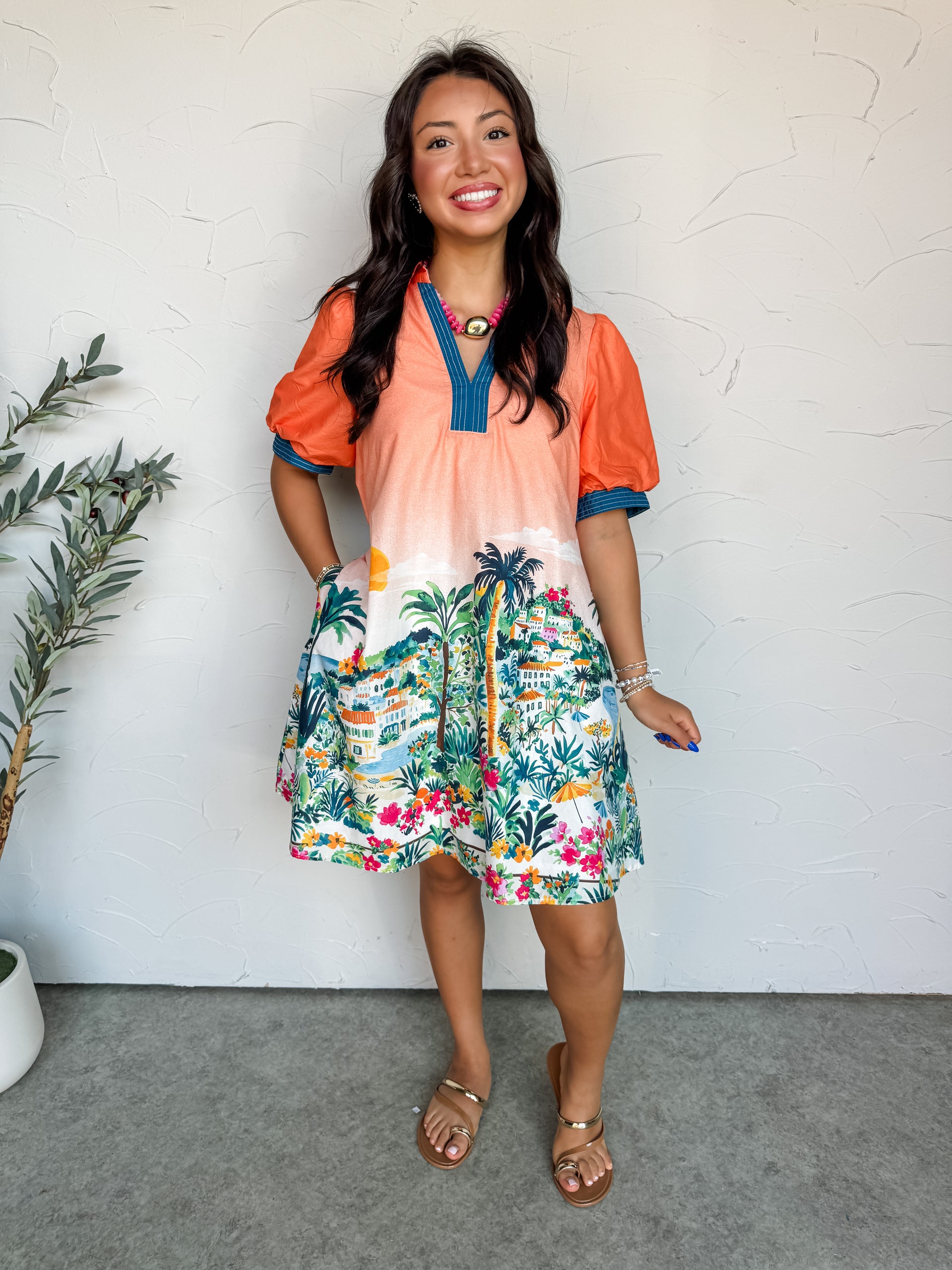 A full-length photograph of a smiling brunette woman posing with one hand in her pocket against a textured white wall. She is wearing a short puff-sleeve shift dress made from 100% cotton fabric featuring an orange-to-white ombre top and a detailed tropical coastal village print along the skirt, styled with contrast navy stitched trim on the V-neckline and sleeve cuffs, and flat gold toe-loop sandals.