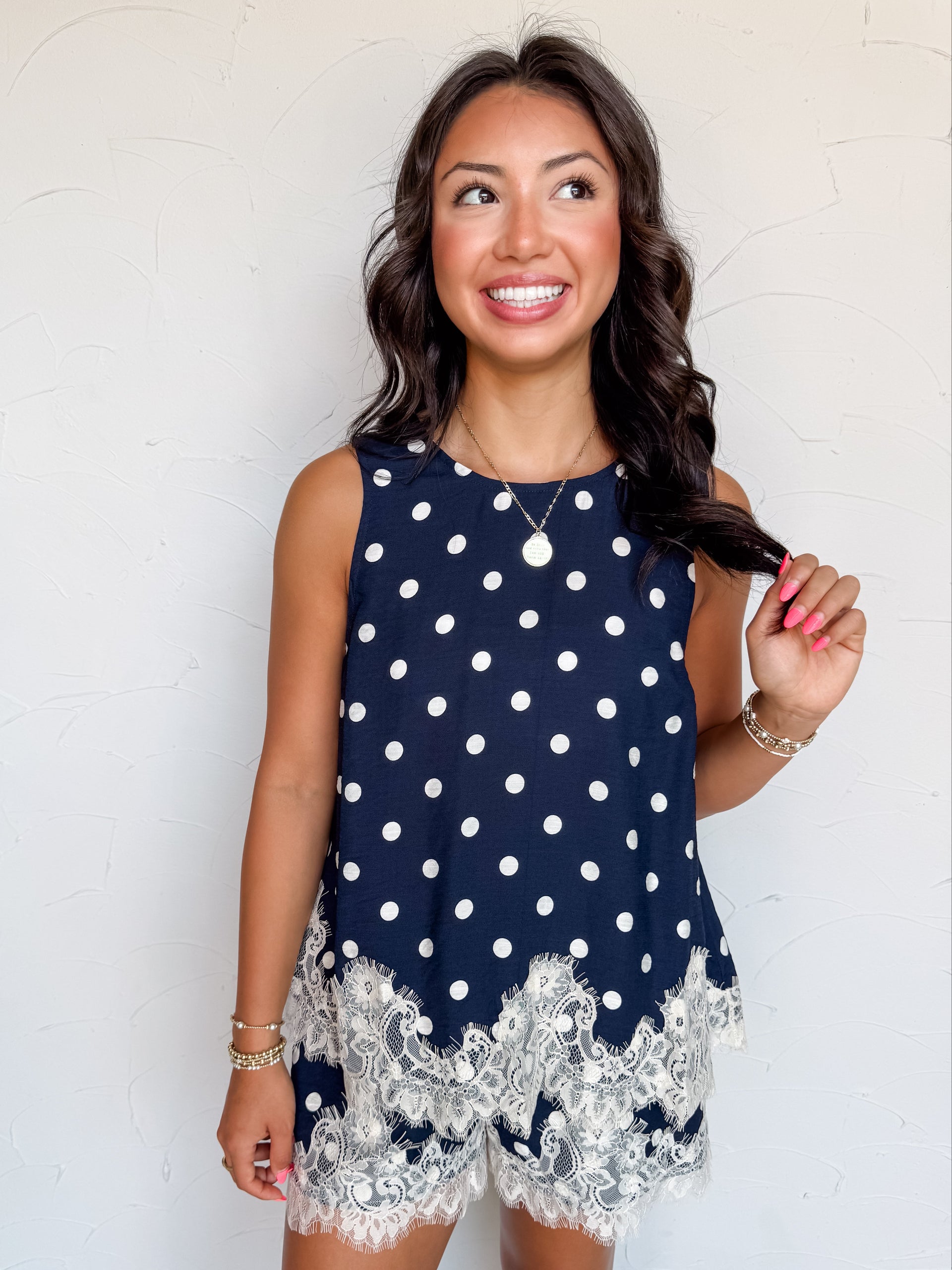 A waist-up photograph of a smiling woman with dark wavy hair holding a strand of her hair against a textured white wall. She is wearing a sleeveless navy blue top with a white polka dot print, made from an 81% viscose and 19% nylon blend, featuring a white eyelash lace trim along the hem, paired with matching polka dot lace-trimmed shorts and styled with a gold coin pendant necklace and gold beaded bracelets.
