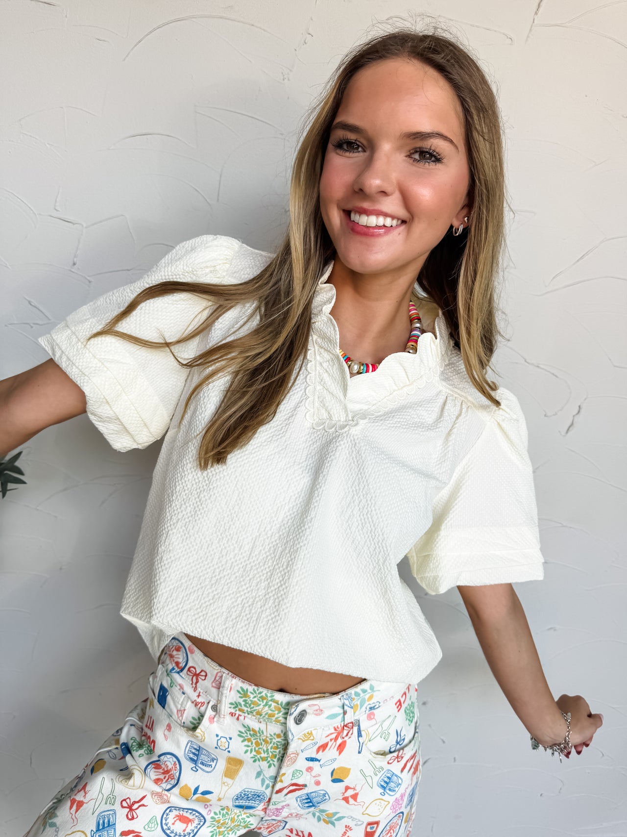 A mid-shot photograph of a smiling blonde woman posing dynamically against a textured white wall. She is wearing a cream-colored short-sleeve top made from a textured 100% polyester fabric, featuring a ruffled split neckline with lace trim detailing and short puff sleeves, styled over white denim pants decorated with colorful Mediterranean illustrations.