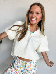 A mid-shot photograph of a smiling blonde woman posing dynamically against a textured white wall. She is wearing a cream-colored short-sleeve top made from a textured 100% polyester fabric, featuring a ruffled split neckline with lace trim detailing and short puff sleeves, styled over white denim pants decorated with colorful Mediterranean illustrations.