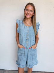 A knee-up photograph of a smiling young woman with long, straight blonde-highlighted hair placing her hands in her pockets against a textured white wall. She is wearing a light-wash denim romper made from 100% cotton, featuring flutter cap sleeves, a scoop neckline, a button-down placket with round wooden buttons, two front patch pockets, and ruffled leg cuffs, styled with a chunky gold chain heart necklace and delicate silver and pearl bracelets.