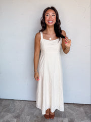 A full-length photograph of a smiling woman with dark wavy hair touching a strand of her hair and looking upward against a textured white wall. She is wearing a sleeveless cream midi dress made from a 64% polyester and 36% rayon blend, featuring wide shoulder straps, a scoop neckline, a fitted bodice with subtle tonal jacquard floral details, and a flowing flared skirt, styled with cognac brown leather shoes, a gold coin pendant necklace, and gold beaded bracelets.