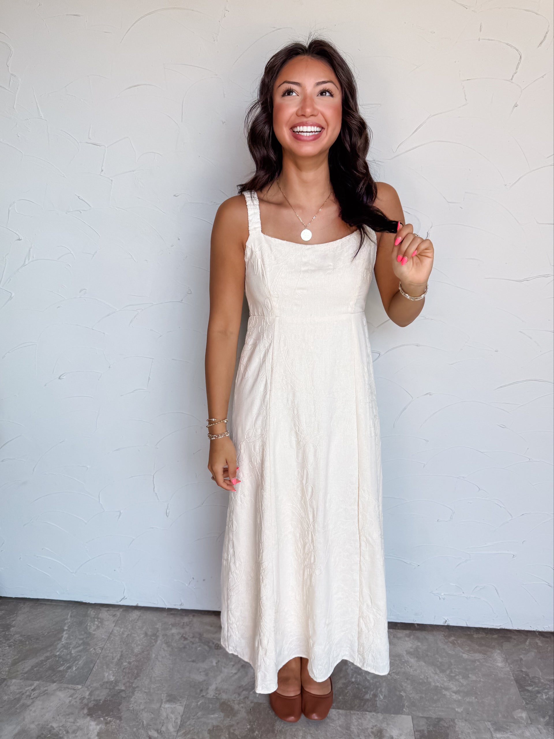 A full-length photograph of a smiling woman with dark wavy hair touching a strand of her hair and looking upward against a textured white wall. She is wearing a sleeveless cream midi dress made from a 64% polyester and 36% rayon blend, featuring wide shoulder straps, a scoop neckline, a fitted bodice with subtle tonal jacquard floral details, and a flowing flared skirt, styled with cognac brown leather shoes, a gold coin pendant necklace, and gold beaded bracelets.