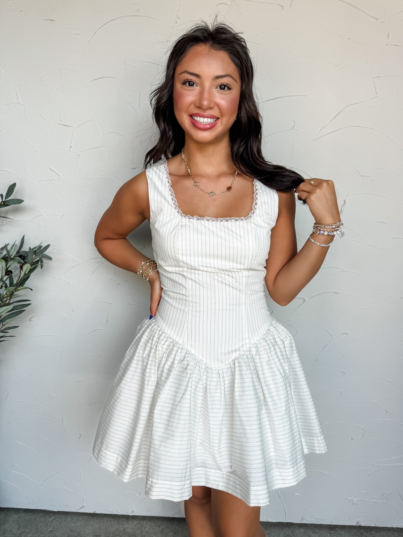 A mid-shot photograph of a smiling brunette woman posing with one hand on her hip and the other touching her hair against a textured white wall. She is wearing a sleeveless white mini dress made from an 85% rayon and 15% nylon blend fabric featuring faint blue pinstripes, designed with delicate white lace trim on the neckline and a gathered drop-waist flared skirt.