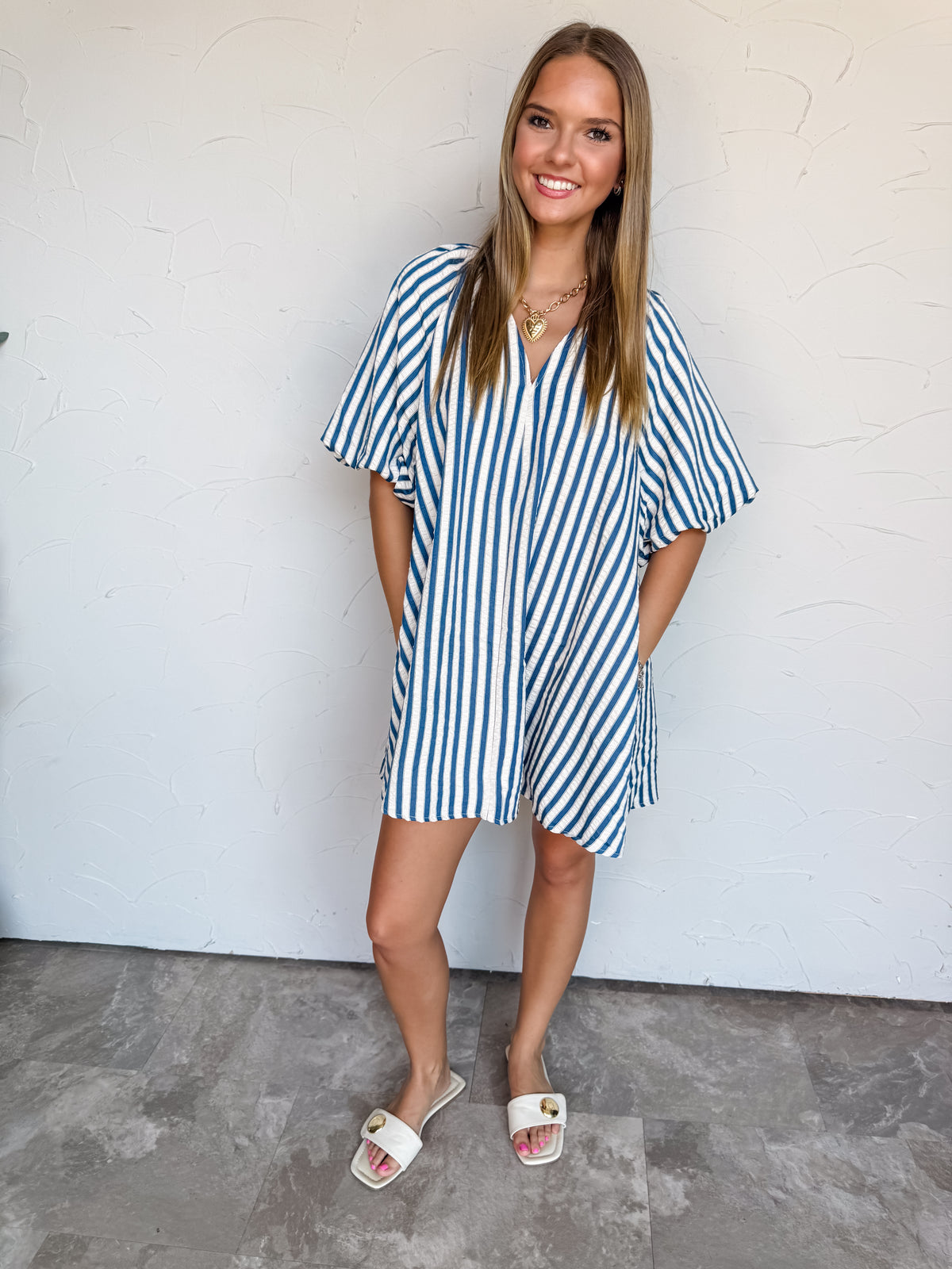 A full-length photograph of a smiling young woman with long, straight blonde-highlighted hair posing playfully on one leg against a textured white wall. She is wearing an oversized short-sleeved mini dress made from 100% polyester textured fabric, featuring bright blue and white vertical stripes, wide elbow-length sleeves, and a open V-neckline, paired with cream open-toe slide heels and styled with a chunky gold chain heart necklace and silver charm bracelets.