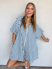 A full-length photograph of a smiling young woman with long, straight blonde-highlighted hair posing playfully on one leg against a textured white wall. She is wearing an oversized short-sleeved mini dress made from 100% polyester textured fabric, featuring bright blue and white vertical stripes, wide elbow-length sleeves, and a open V-neckline, paired with cream open-toe slide heels and styled with a chunky gold chain heart necklace and silver charm bracelets.