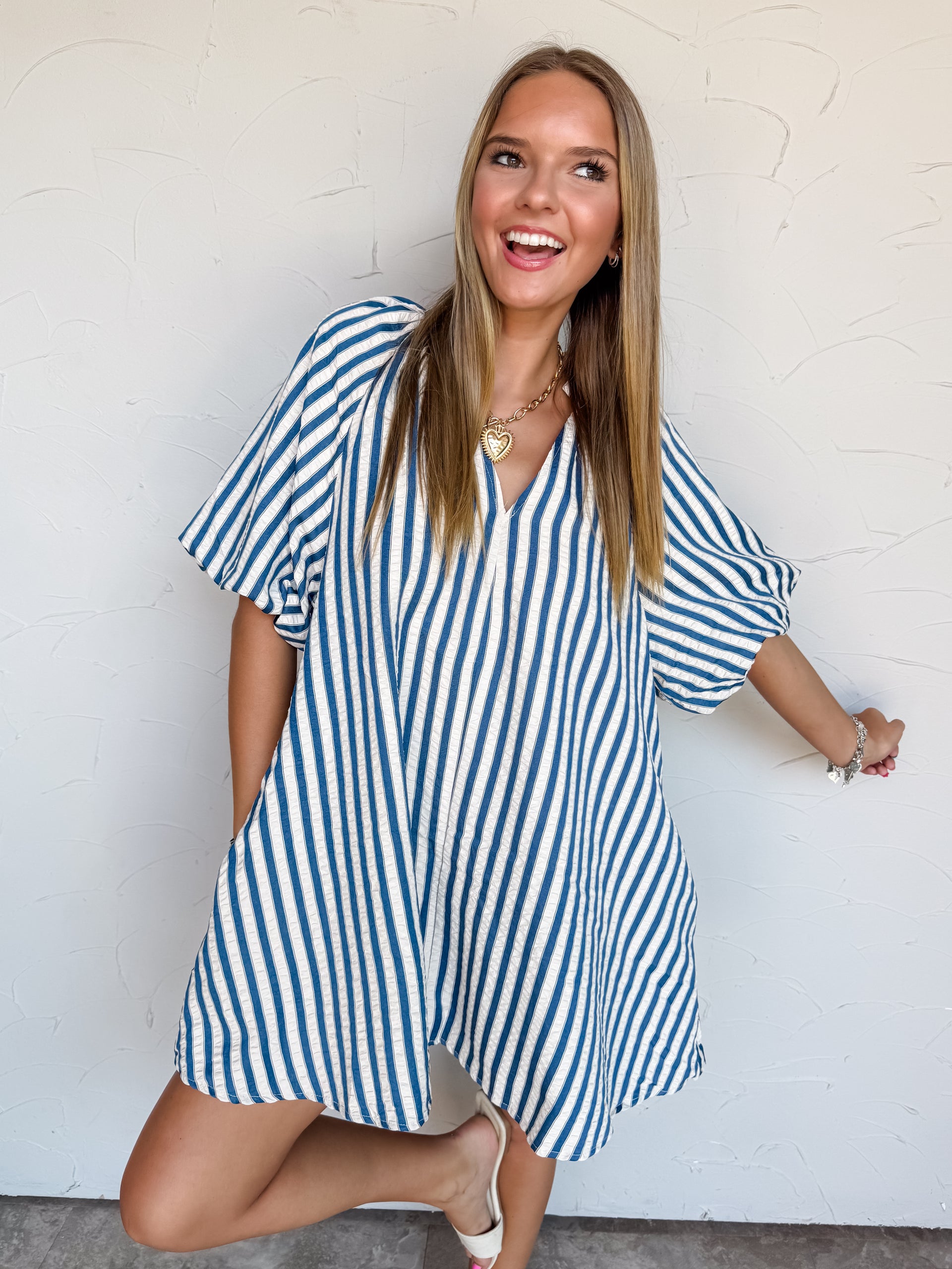 A full-length photograph of a smiling young woman with long, straight blonde-highlighted hair posing playfully on one leg against a textured white wall. She is wearing an oversized short-sleeved mini dress made from 100% polyester textured fabric, featuring bright blue and white vertical stripes, wide elbow-length sleeves, and a open V-neckline, paired with cream open-toe slide heels and styled with a chunky gold chain heart necklace and silver charm bracelets.