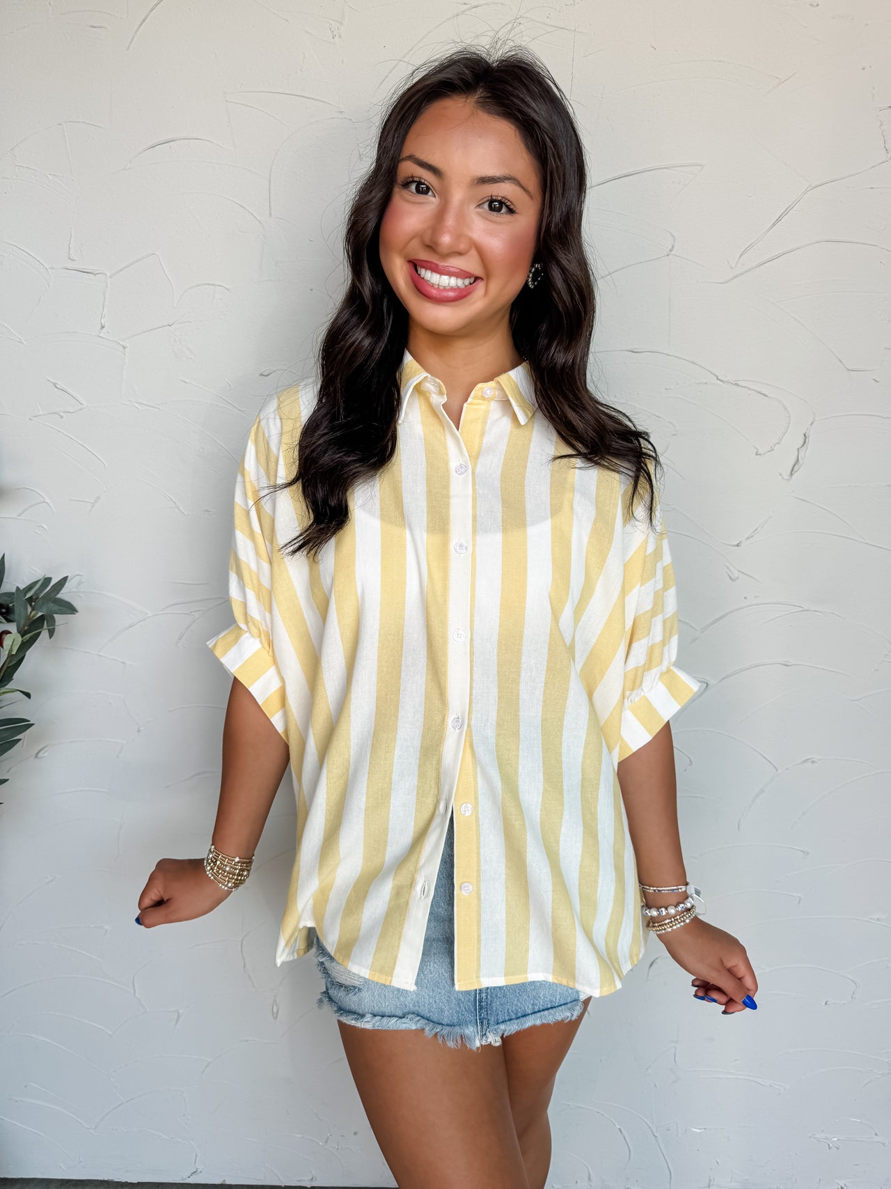 A mid-shot photograph of a smiling brunette woman posing against a textured white wall. She is wearing a relaxed, short-sleeve button-down top made from a 70% cotton and 30% polyester blend fabric featuring yellow and white vertical stripes, styled with white buttons and cuffed sleeves over distressed light-wash denim shorts.