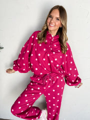 Model wearing the Just Between Us Pink Polka Dot Lounge Set, made from 50% cotton and 50% polyester. The bright pink set features light pink polka dots, a long-sleeve top with a pointed collar, partial snap-front placket, and ribbed cuffs and hem, paired with matching full-length pants. Styled with pink sneakers and layered jewelry against a white brick wall.