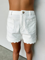 A close-up photograph focusing from the waist down of a woman wearing high-waisted white denim shorts made from a 97% cotton and 3% spandex blend fabric against a textured white wall. The shorts feature unique scalloped borders on the front patch pockets and along the bottom leg hems, complete with a brown horn-style button at the waist closure.