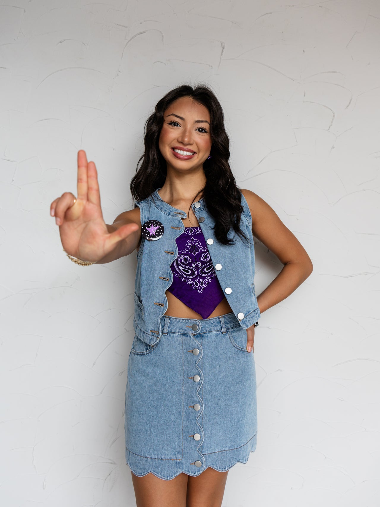 A mid-shot photograph of a smiling young woman with long dark wavy hair gesturing toward the camera with one hand while posing against a textured white wall. She is wearing a matching two-piece light wash denim set made from 100% cotton fabric, featuring a sleeveless snap-button vest accented with a pin and a matching button-front mini skirt with a scalloped hemline, styled over a purple paisley bandana tube top.