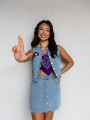 A mid-shot photograph of a smiling young woman with long dark wavy hair gesturing toward the camera with one hand while posing against a textured white wall. She is wearing a matching two-piece light wash denim set made from 100% cotton fabric, featuring a sleeveless snap-button vest accented with a pin and a matching button-front mini skirt with a scalloped hemline, styled over a purple paisley bandana tube top.