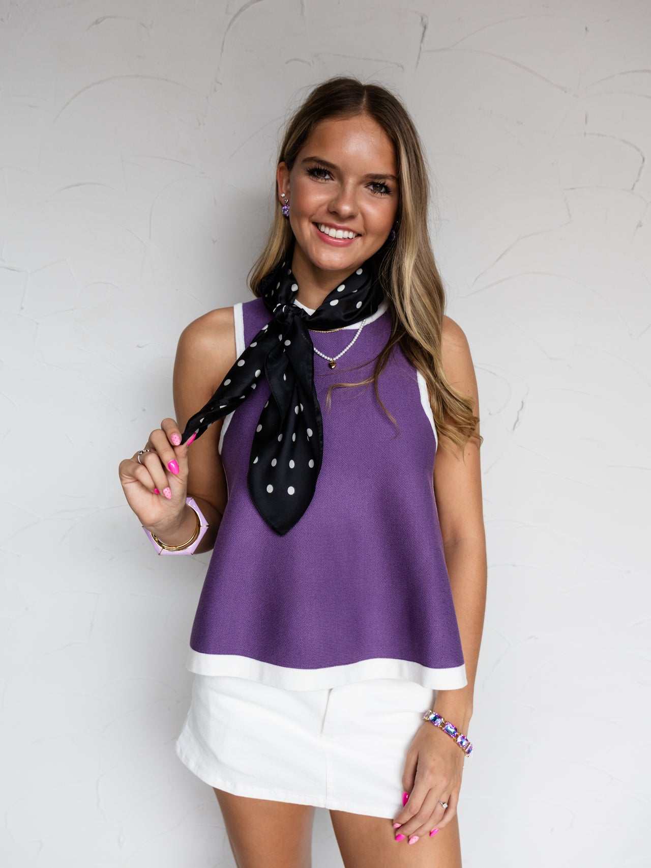 A mid-shot photograph of a smiling young woman with wavy light brown hair posing against a textured white wall while holding a corner of her neck scarf. She is wearing a purple sleeveless knit tank top made from a 100% viscose fabric, featuring a white contrast trim along the neckline and hemline, styled with a white mini skirt, a black-and-white polka dot neck scarf, and bracelets.