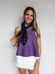 A mid-shot photograph of a smiling young woman with wavy light brown hair posing against a textured white wall while holding a corner of her neck scarf. She is wearing a purple sleeveless knit tank top made from a 100% viscose fabric, featuring a white contrast trim along the neckline and hemline, styled with a white mini skirt, a black-and-white polka dot neck scarf, and bracelets.
