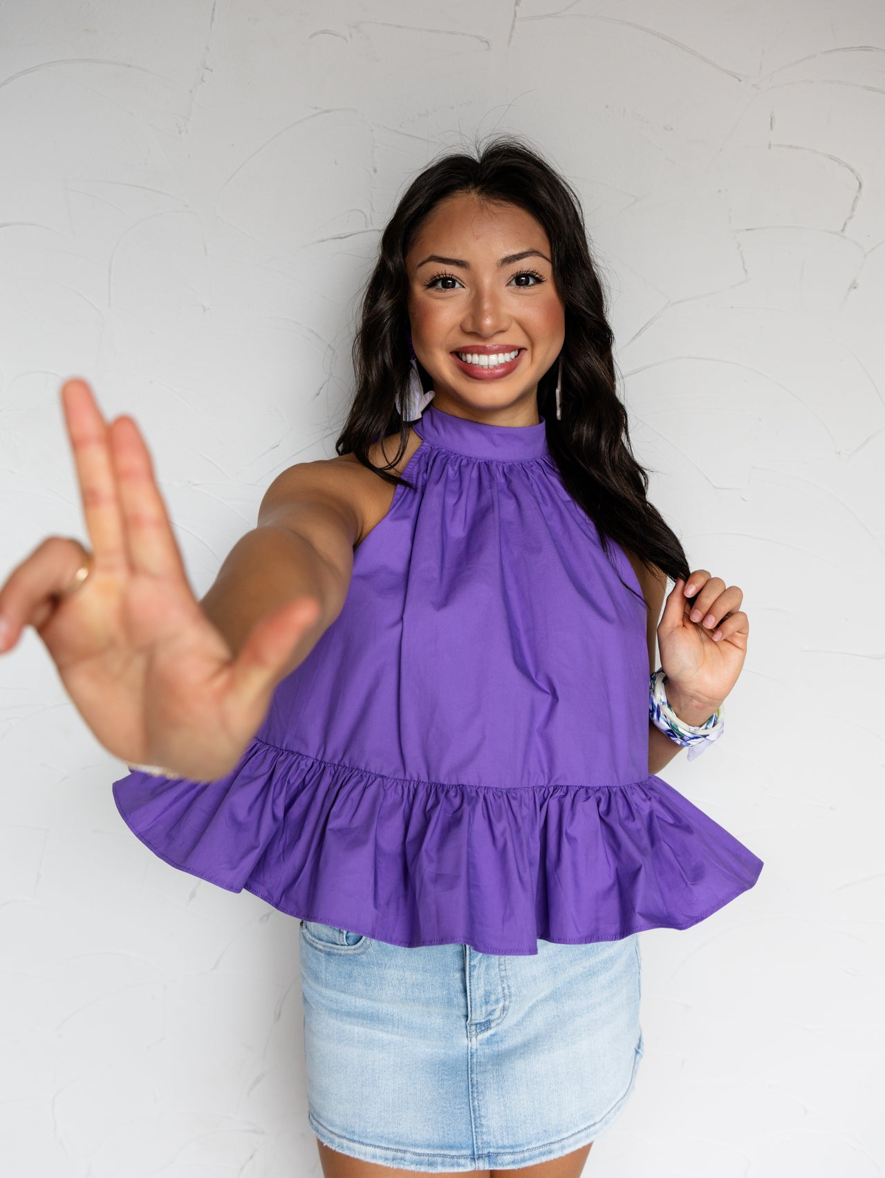 A mid-shot photograph of a smiling young woman with long dark wavy hair gesturing toward the camera with one hand while posing against a textured white wall. She is wearing a vibrant purple halter tank top made from a 100% cotton fabric, featuring a high collar and a flared ruffle hemline, styled with a light wash denim skirt and a patterned bracelet.
