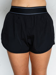 A close-up photograph focusing from the waist down of a person standing against a textured white wall. They are wearing black athletic running shorts made from an 86% nylon and 14% spandex blend fabric, featuring a high-rise elastic waistband with coordinating thin white lines and curved dolphin-style hems.