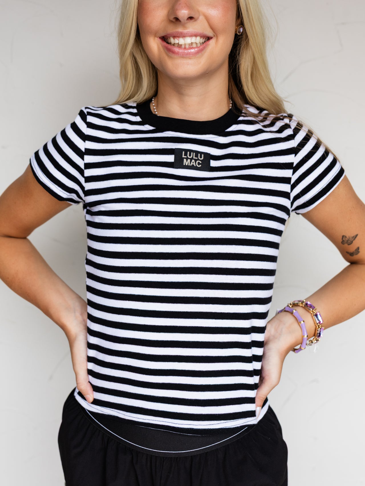 A close-up photograph focusing from the waist to the neck of a smiling blonde woman posing with both hands on her hips against a textured white wall. She is wearing a fitted black and white horizontally striped baby tee made from a 97% cotton and 3% spandex blend fabric, featuring a solid black crew neckline and a small rectangular black patch at the center chest that reads "LULU MAC".