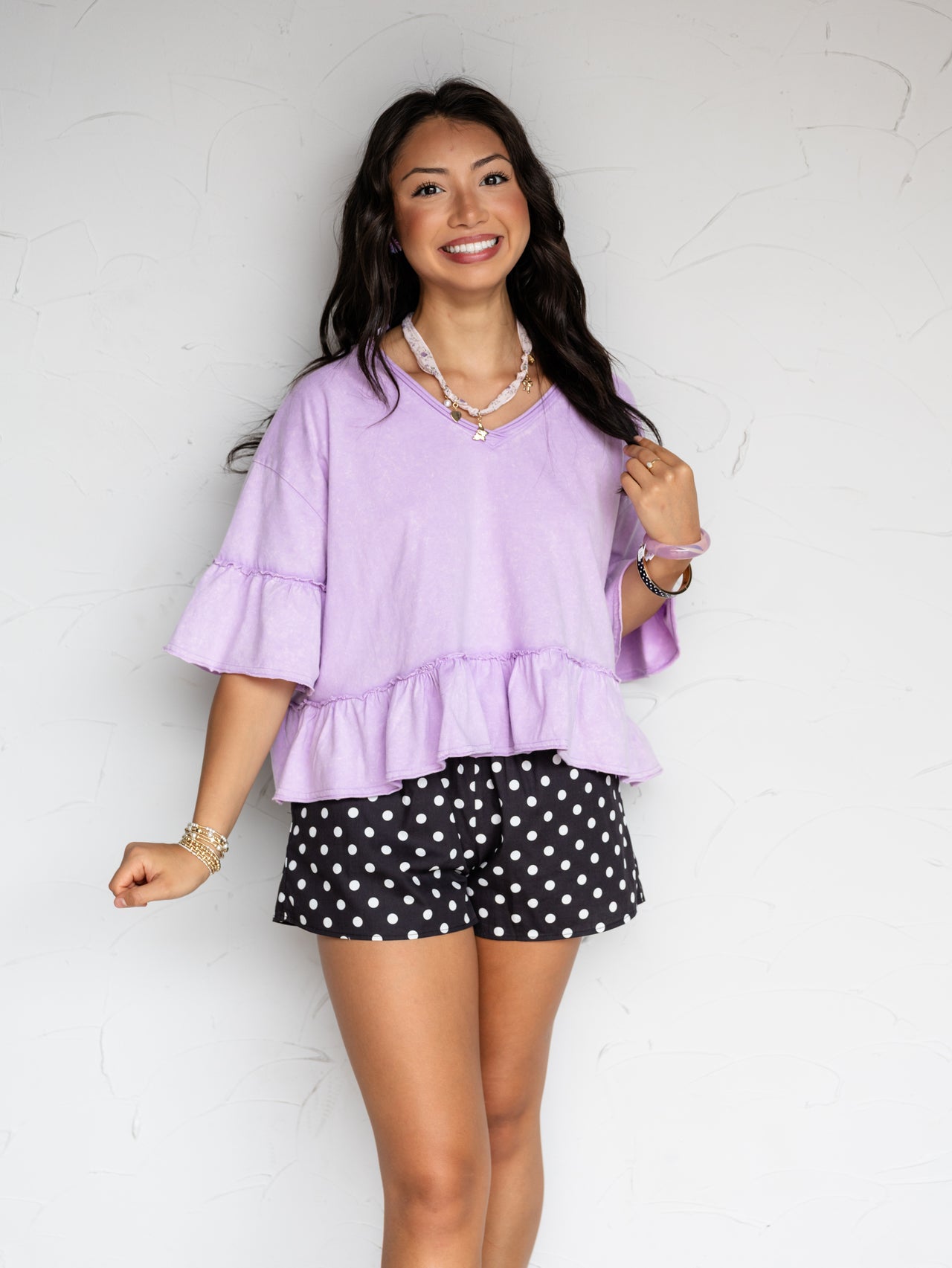 A mid-shot photograph of a smiling young woman with long dark wavy hair posing while holding a strand of hair against a textured white wall. She is wearing a relaxed lavender V-neck top made from a 100% cotton fabric, featuring tiered ruffles on the short sleeves and a ruffled peplum hemline, styled with black and white polka dot boxer shorts, a charm necklace, and stacked bracelets.