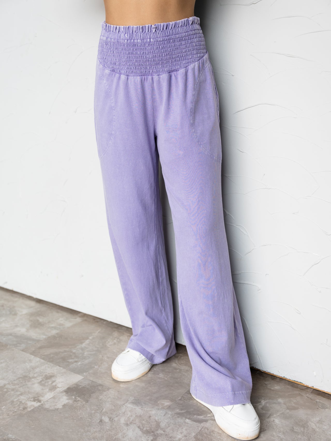 A close-up photograph focusing from the waist down of a person standing against a textured white wall. They are wearing relaxed beige wide-leg pants made from 100% cotton fabric, featuring a high-rise, thick smocked elastic waistband and subtle side pockets, styled with neutral-colored sneakers.