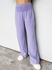 A close-up photograph focusing from the waist down of a person standing against a textured white wall. They are wearing relaxed beige wide-leg pants made from 100% cotton fabric, featuring a high-rise, thick smocked elastic waistband and subtle side pockets, styled with neutral-colored sneakers.