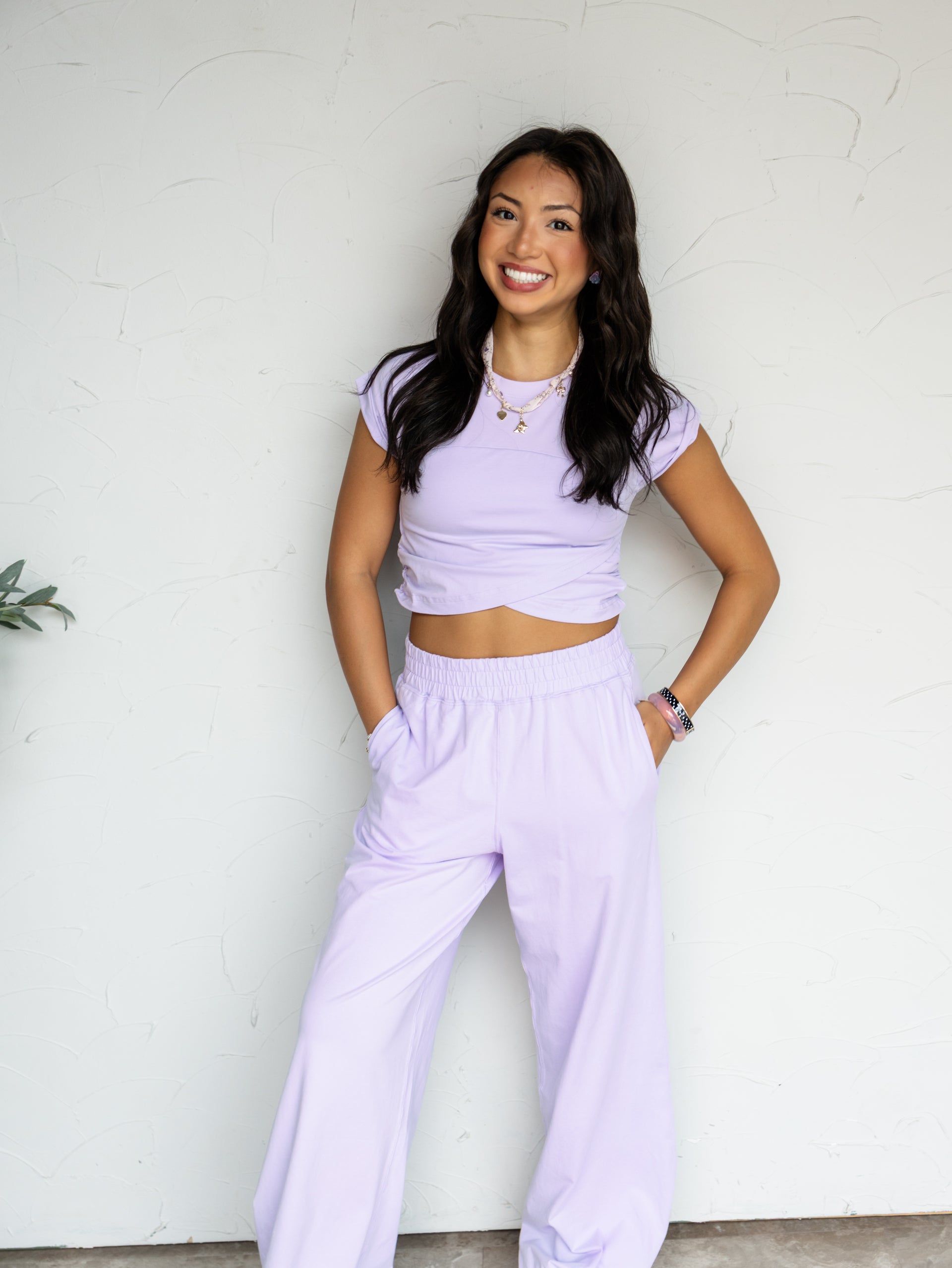 A full-length photograph of a smiling young woman with long dark wavy hair posing with both hands in her pants pockets against a textured white wall. She is wearing a matching two-piece lavender set made from a 95% cotton and 5% spandex blend fabric, featuring a crewneck crop top with capped sleeves and an overlapping tulip hemline, paired with matching high-waisted wide-leg pants, styled with a charm necklace and stacked bracelets.