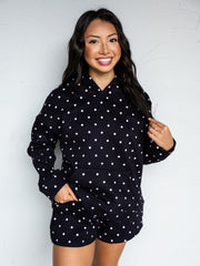 A mid-shot photograph of a smiling young woman with long dark wavy hair posing while holding a strand of her hair against a textured white wall. She is wearing a matching two-piece black lounge set covered in a white polka dot pattern, made from a 50% cotton and 50% polyester blend fabric, featuring a relaxed pullover hoodie with a front kangaroo pocket and matching sweat shorts.