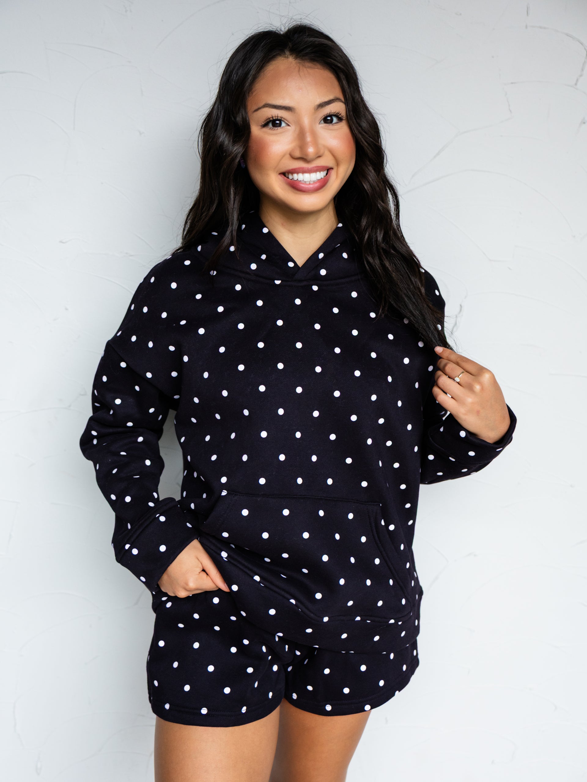 A mid-shot photograph of a smiling young woman with long dark wavy hair posing while holding a strand of her hair against a textured white wall. She is wearing a matching two-piece black lounge set covered in a white polka dot pattern, made from a 50% cotton and 50% polyester blend fabric, featuring a relaxed pullover hoodie with a front kangaroo pocket and matching sweat shorts.