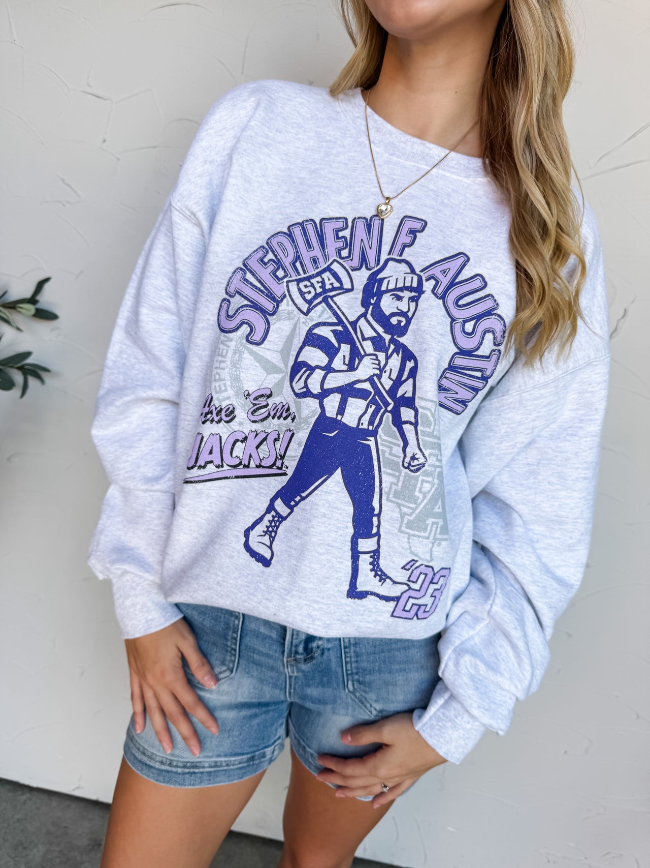 Woman wearing a gray sweatshirt with a graphic design and a purple cap, standing against a light-colored wall.