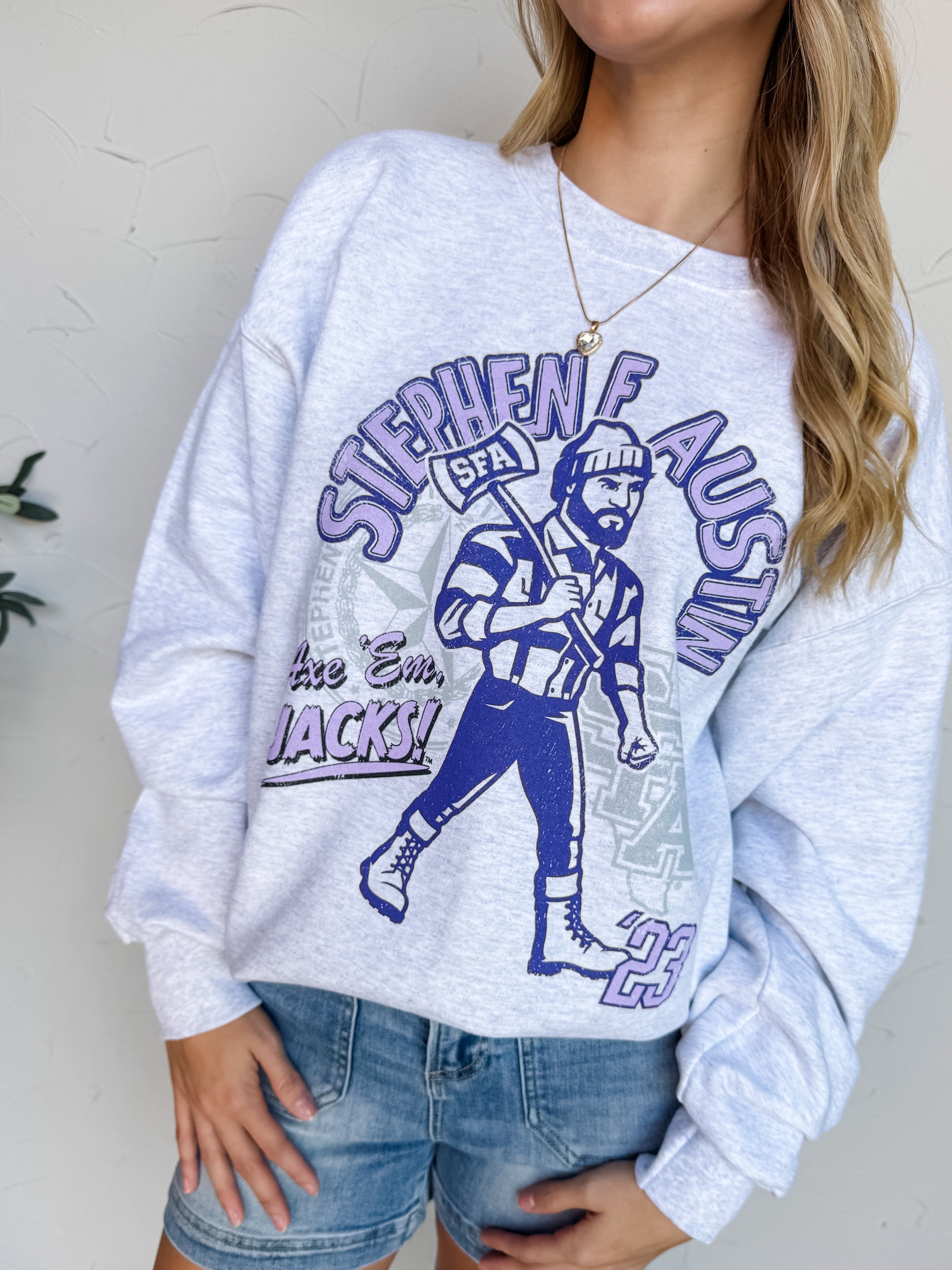 Woman wearing a gray sweatshirt with a graphic design and a purple cap, standing against a light-colored wall.