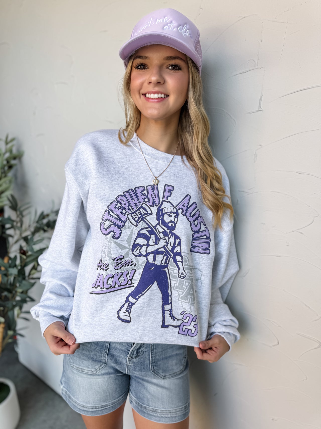 Woman wearing a gray sweatshirt with a graphic design and a purple cap, standing against a light-colored wall.