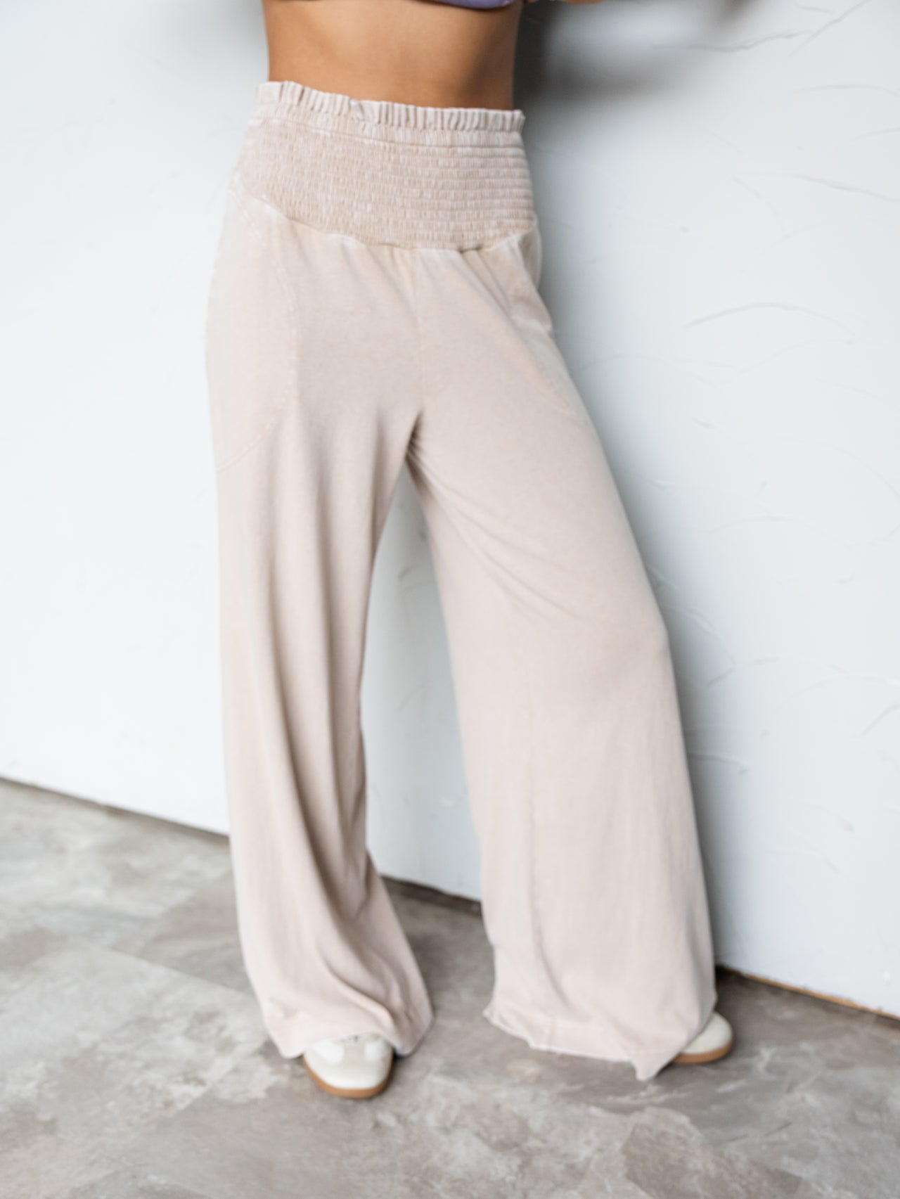 A close-up photograph focusing from the waist down of a person standing against a textured white wall. They are wearing relaxed beige wide-leg pants made from 100% cotton fabric, featuring a high-rise, thick smocked elastic waistband and subtle side pockets, styled with neutral-colored sneakers.
