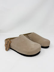 A pair of taupe suede slip-on clogs with black soles, shown on a clean white studio background. A tan leather tag on the side of the front clog reads 'PROPERLY LOVED'.