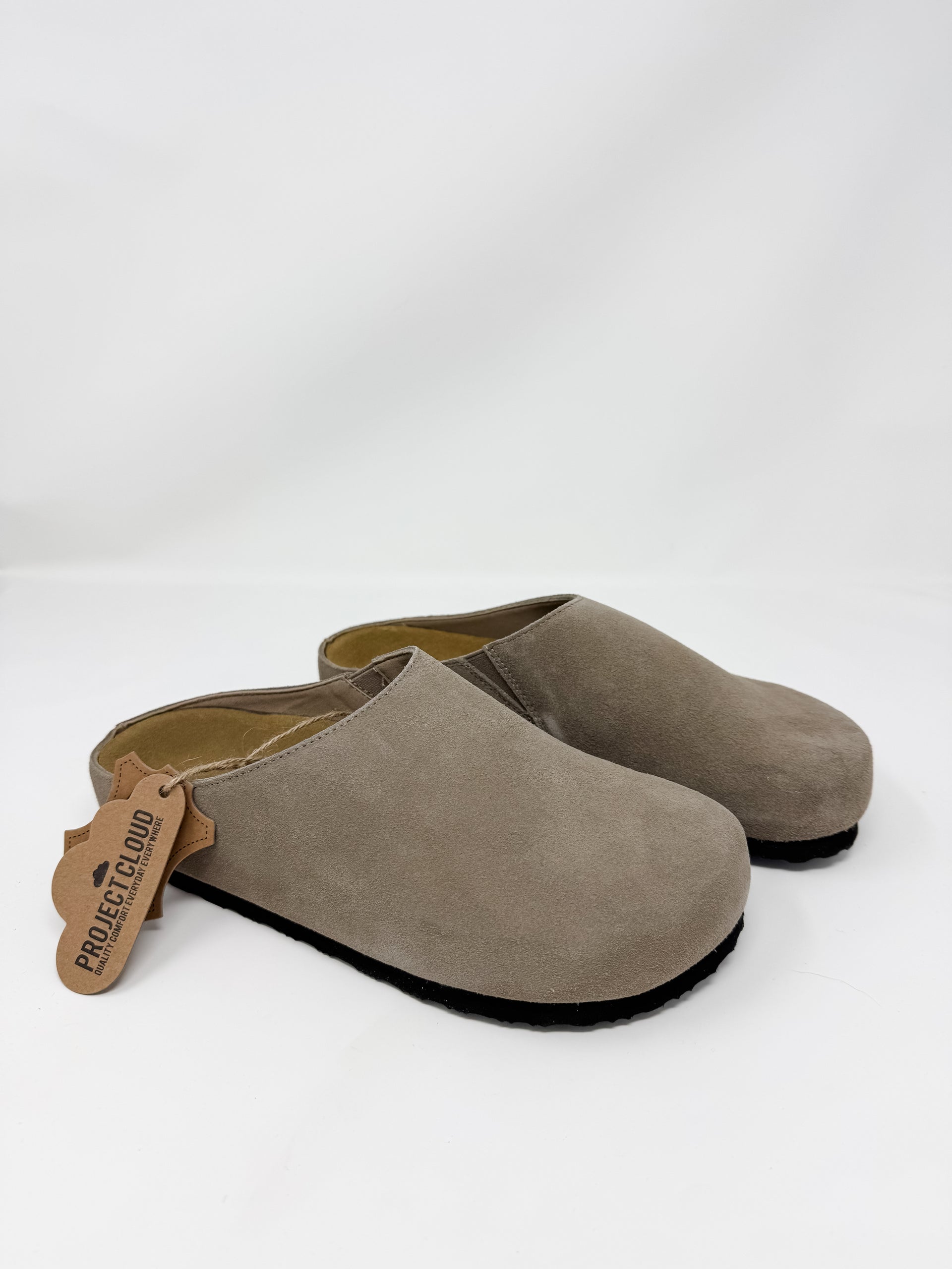 A pair of taupe-brown suede slip-on clogs with black soles, shown on a white studio background. A brown leather brand tag reading 'PROLEGGERO' is attached to the front shoe.