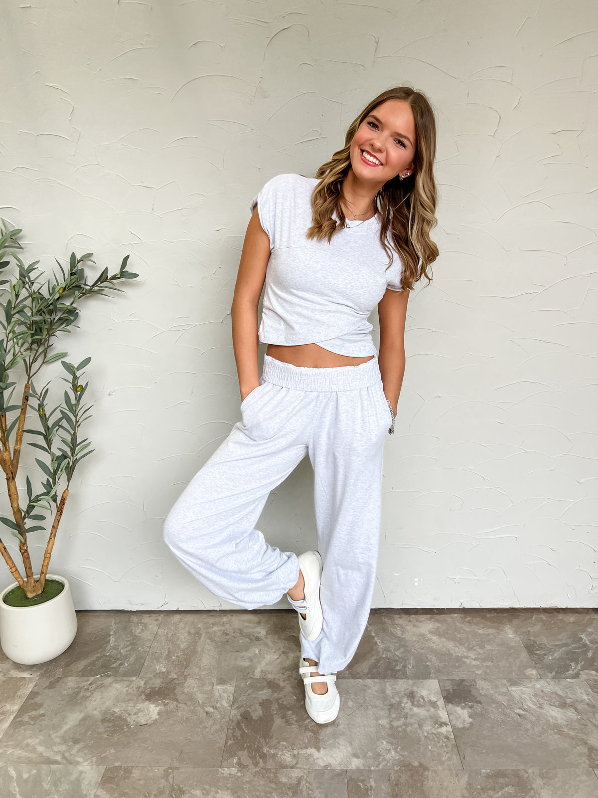 Catie modeling the Rae Mode Feeling Sporty Crossover Set in heather grey, made from 97% cotton and 3% spandex, featuring a cropped cap-sleeve top with an overlapping crossover hem and matching pants with a gathered waistband and side pockets.