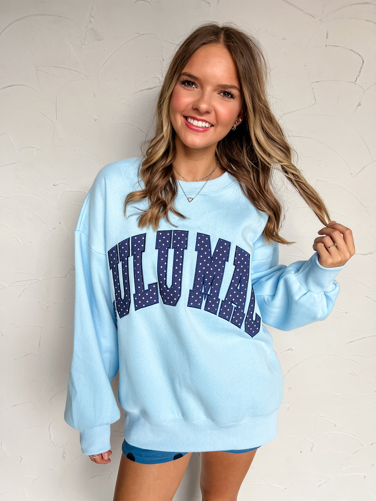 Light blue Lulu Mac crewneck sweatshirt made of 50% cotton and 50% polyester, with navy lettering filled with white polka dots and long sleeves.

