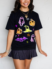 A woman wearing a black short-sleeve graphic T-shirt with colorful Halloween-themed designs including pumpkins, ghosts, a witch hat, and bats, paired with a dark polka-dot skirt against a light textured wall.