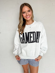 A smiling woman wearing a light heather gray crewneck sweatshirt with 'GAMEDAY' printed across the chest in black varsity-style letters, paired with blue and white striped shorts, standing against a textured off-white wall.Fabric: 52% polyester, 48% cotton
